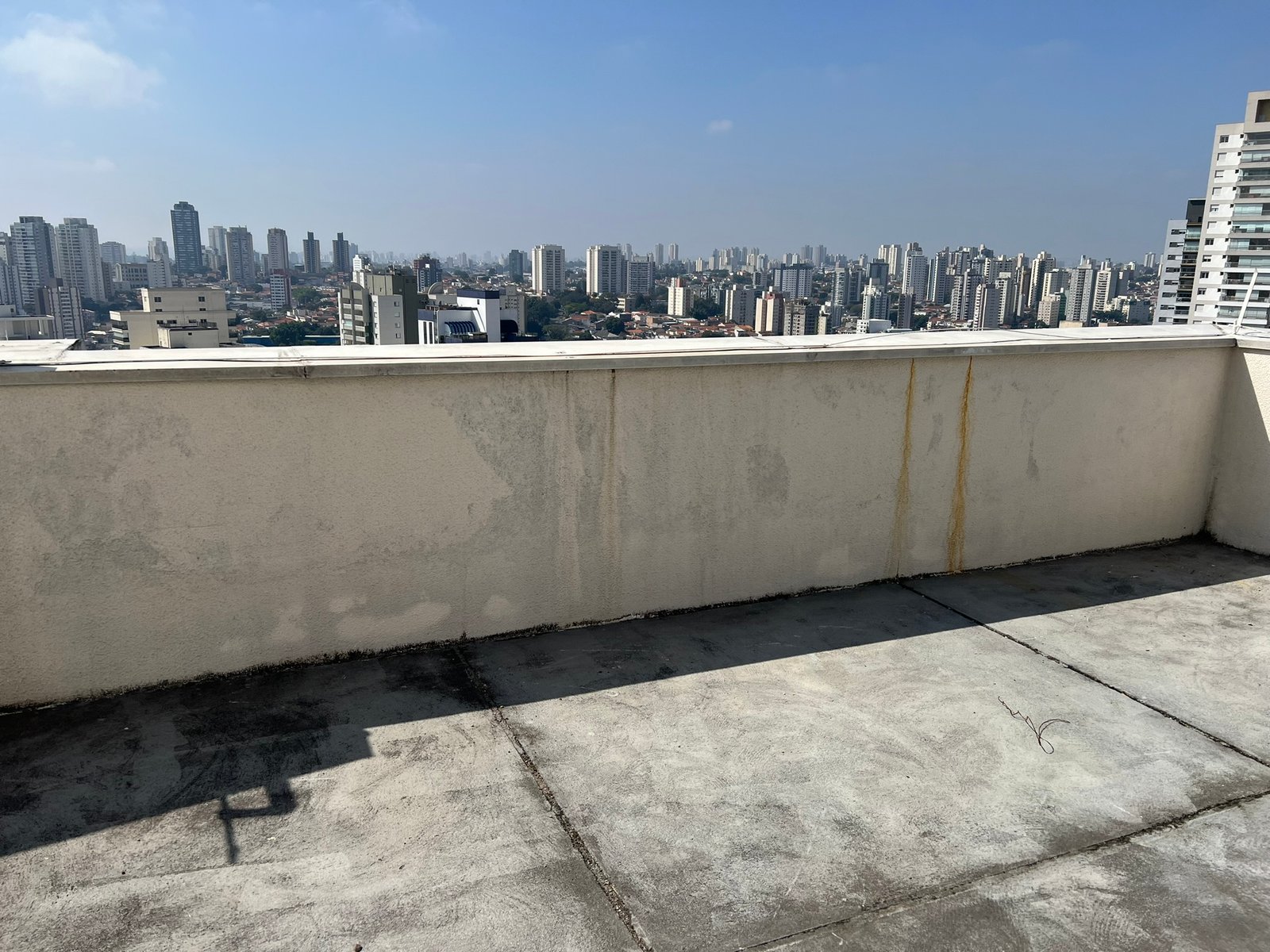 Cobertura de edifício com piso de concreto, parapeito com manchas ferrosas e vista urbana panorâmica.