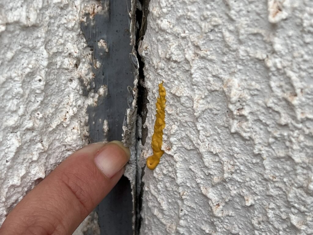 Junta de fachada com selante de vedação amarelo rompido e descolado, dedo tocando o material próximo à fresta entre placas de revestimento.